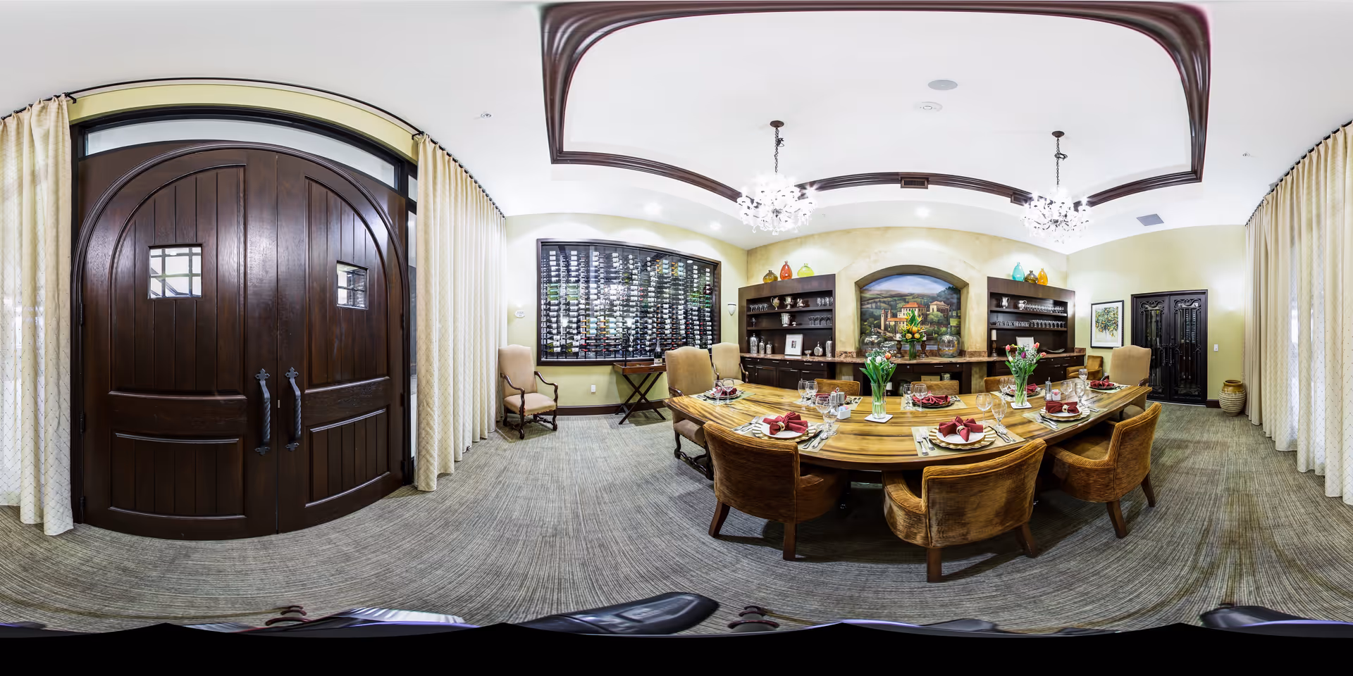 A spacious, elegant dining room with a large wooden table set for eight people with plates, glasses, and red napkins. The room features two large dark wooden double doors, beige curtains covering the windows, and built-in shelves with decorative items and a mural on the back wall. The ceiling has dark wood trim and two chandeliers.