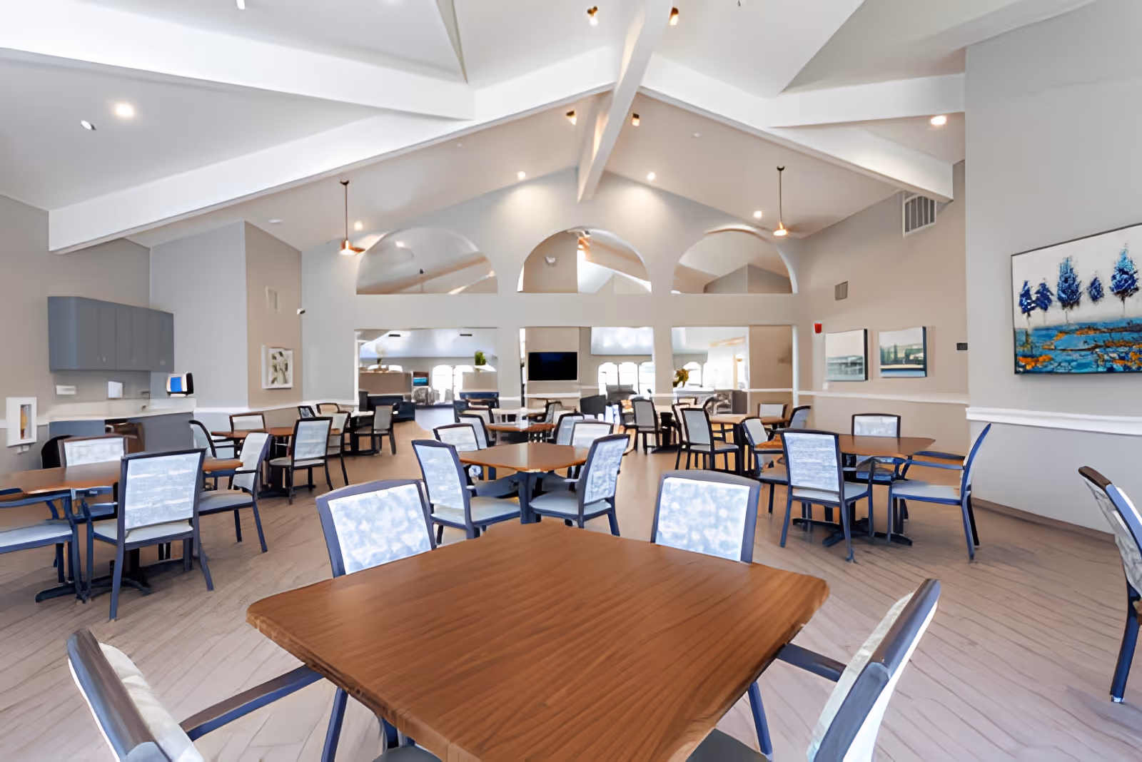 A spacious dining room with multiple wooden tables and cushioned chairs arranged neatly. The room features high vaulted ceilings with exposed beams, pendant lights, and large arched mirrors on the far wall. The walls are painted in neutral tones and decorated with framed artwork. There is a cabinet area on the left side and a television mounted on the far wall.