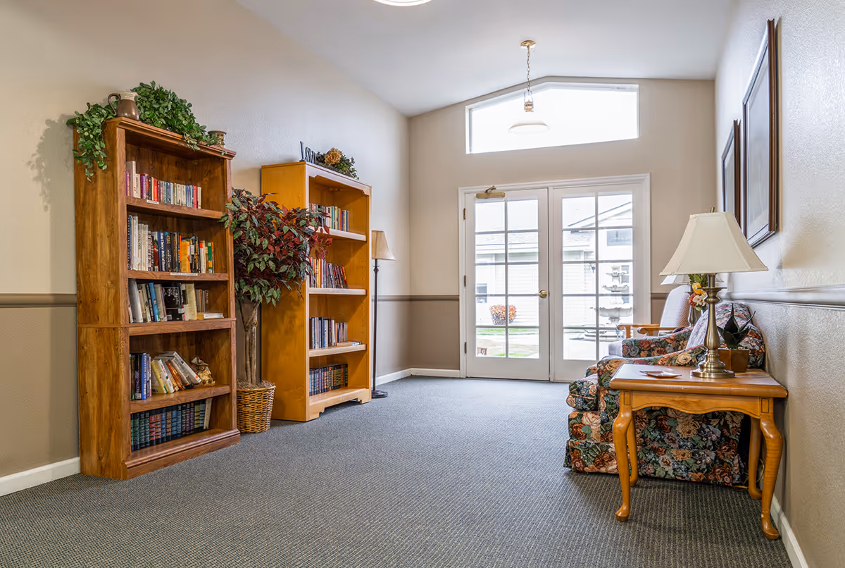 A cozy interior common area with two wooden bookshelves filled with books, a potted plant between them, a floral patterned armchair, a wooden side table with a lamp, and a set of glass double doors leading outside.