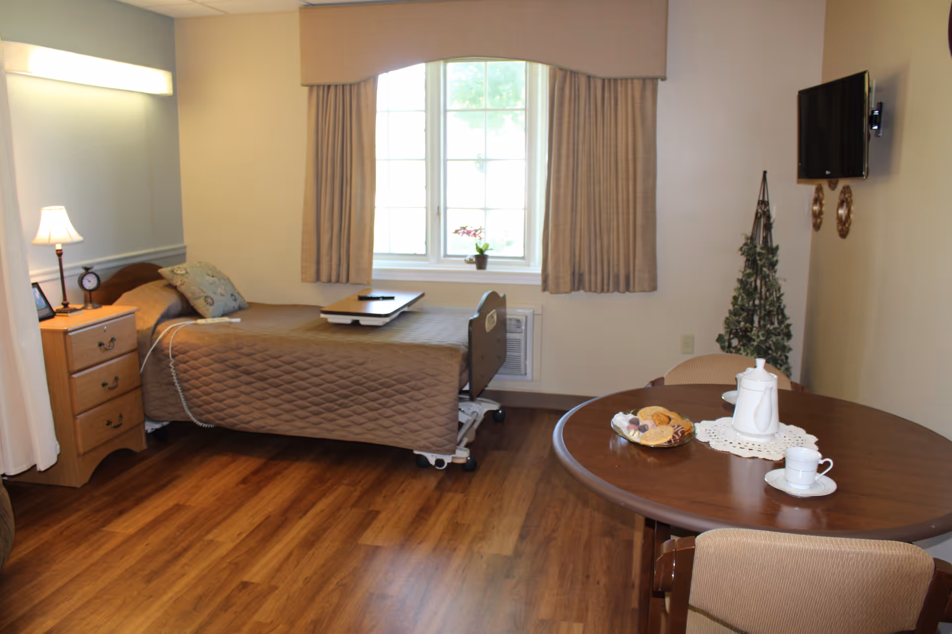 A senior living facility bedroom with a single bed covered in a brown quilt, a bedside table with a lamp and clock, a window with beige curtains, a small artificial Christmas tree, a wall-mounted TV, and a round wooden table set with a white teapot, cup, and a plate of cookies.