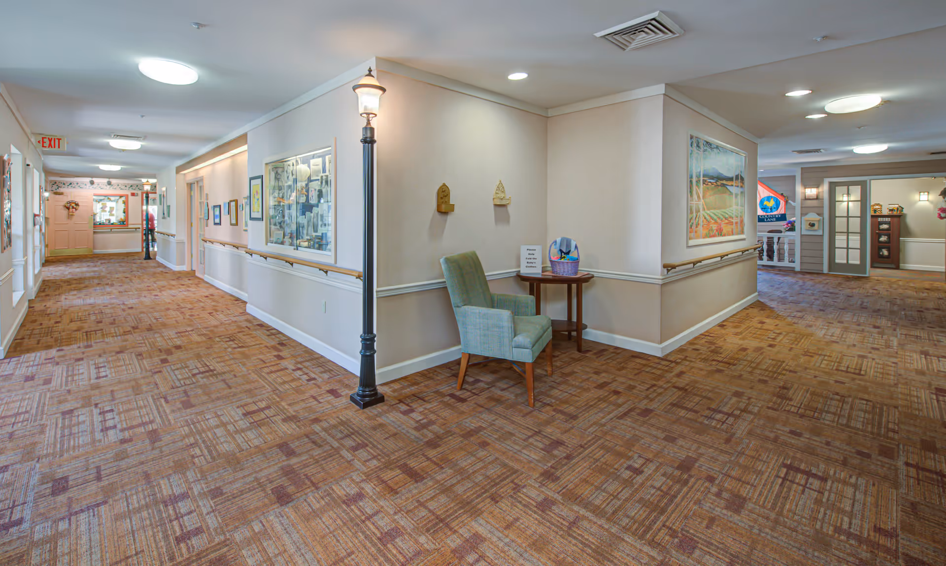 A wide hallway in a senior living facility with patterned carpet, beige walls, and handrails along both sides. There is a green upholstered chair and a small wooden table with a basket and a sign on it near a corner. The hallway is well-lit with ceiling lights and features framed pictures and a display case on the walls. An exit sign is visible at the far end of the hallway.