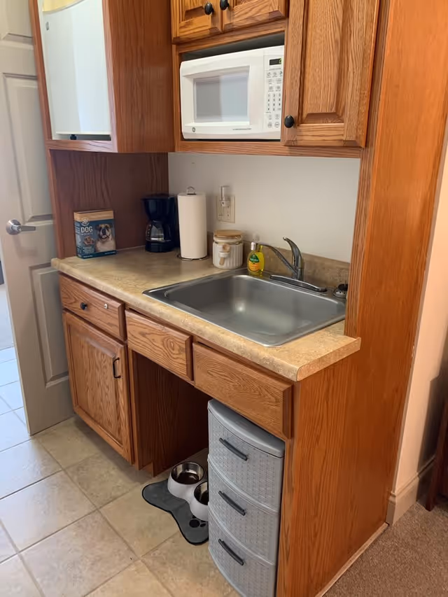 A small kitchen area with wooden cabinets, a countertop with a stainless steel sink, a white microwave mounted above, a coffee maker, paper towel holder, and a container on the counter. Below the counter are drawers and a small plastic storage unit. On the floor, there are two pet bowls on a paw-shaped mat.