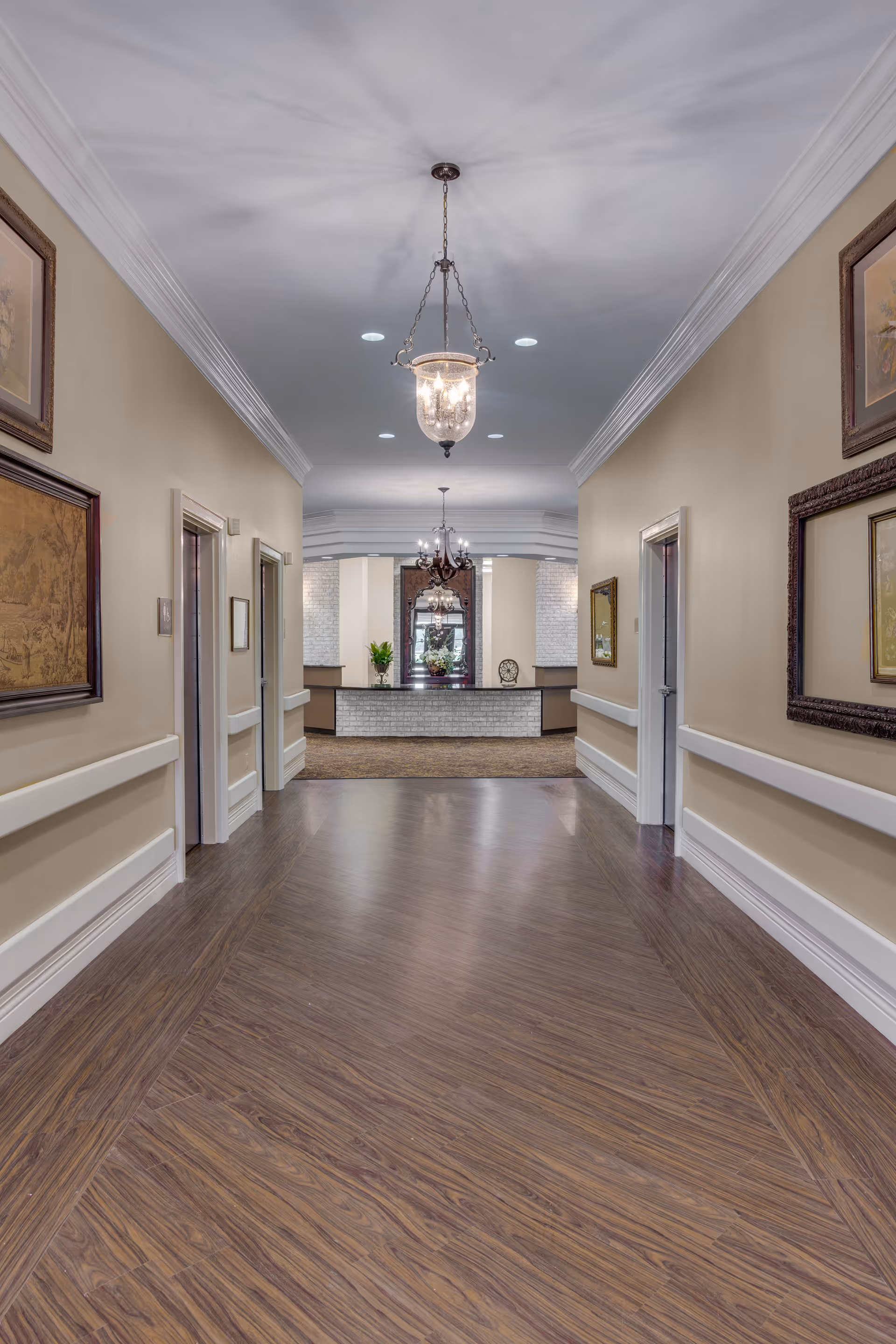 A well-lit interior hallway with wood floors leading to a reception desk and chandeliers.