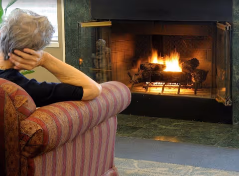 An elderly person with gray hair sitting on a striped upholstered armchair, facing a lit fireplace with a protective screen in a cozy room.