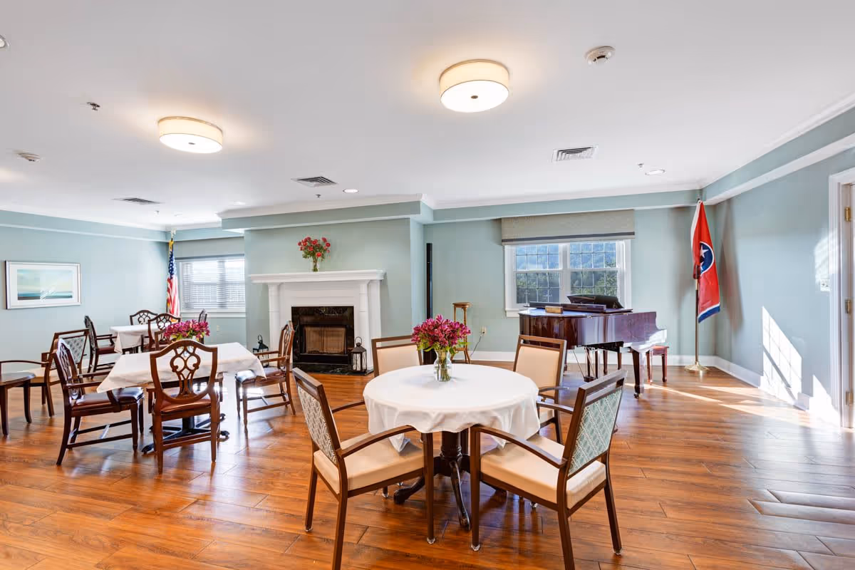 A bright and spacious common room with wooden floors, light blue walls, and ceiling lights. The room features several round and rectangular tables with chairs around them, each table decorated with a vase of flowers. There is a white fireplace with a vase of red flowers on the mantel, an American flag in one corner, and a grand piano near a window with a Tennessee state flag beside it.