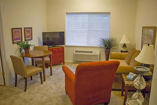 A cozy living room with a red armchair, a beige sofa, a wooden side table with a lamp and tissue box, a small round dining table with two chairs, a flat-screen TV on a red cabinet, a large window with blinds, and decorative plants and framed pictures on the walls.
