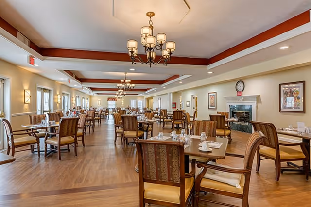 A spacious dining room in a senior living facility with multiple wooden tables and cushioned chairs arranged neatly. The room features warm lighting from chandeliers and wall sconces, a fireplace with a clock above it, framed artwork on the walls, and large windows allowing natural light to enter.