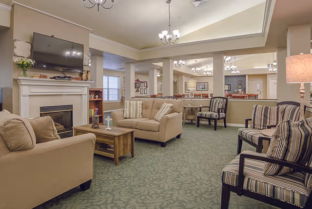 A cozy living room area in a senior living facility with beige sofas, striped armchairs, a wooden coffee table, a fireplace with a mounted flat-screen TV above it, and soft lighting from ceiling chandeliers and a floor lamp.