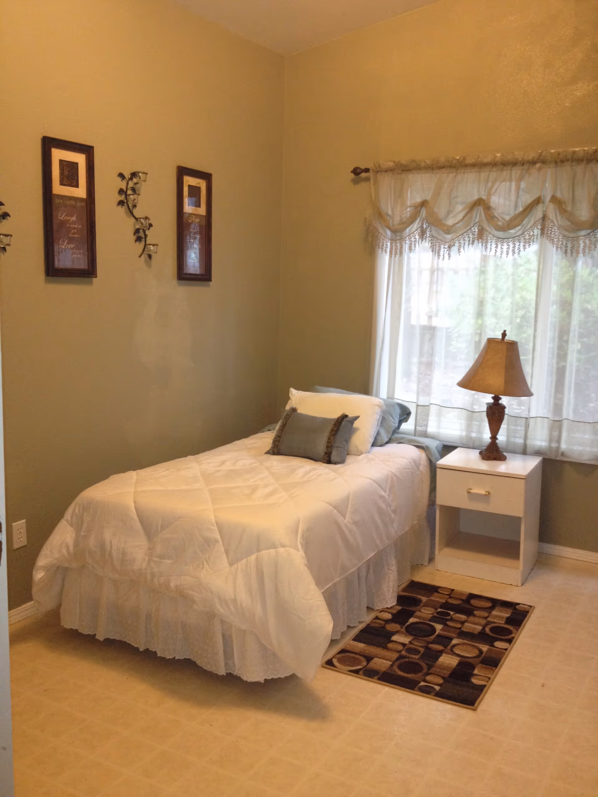 A small bedroom with a single bed dressed in white bedding, a nightstand and lamp by a curtained window and framed wall art.
