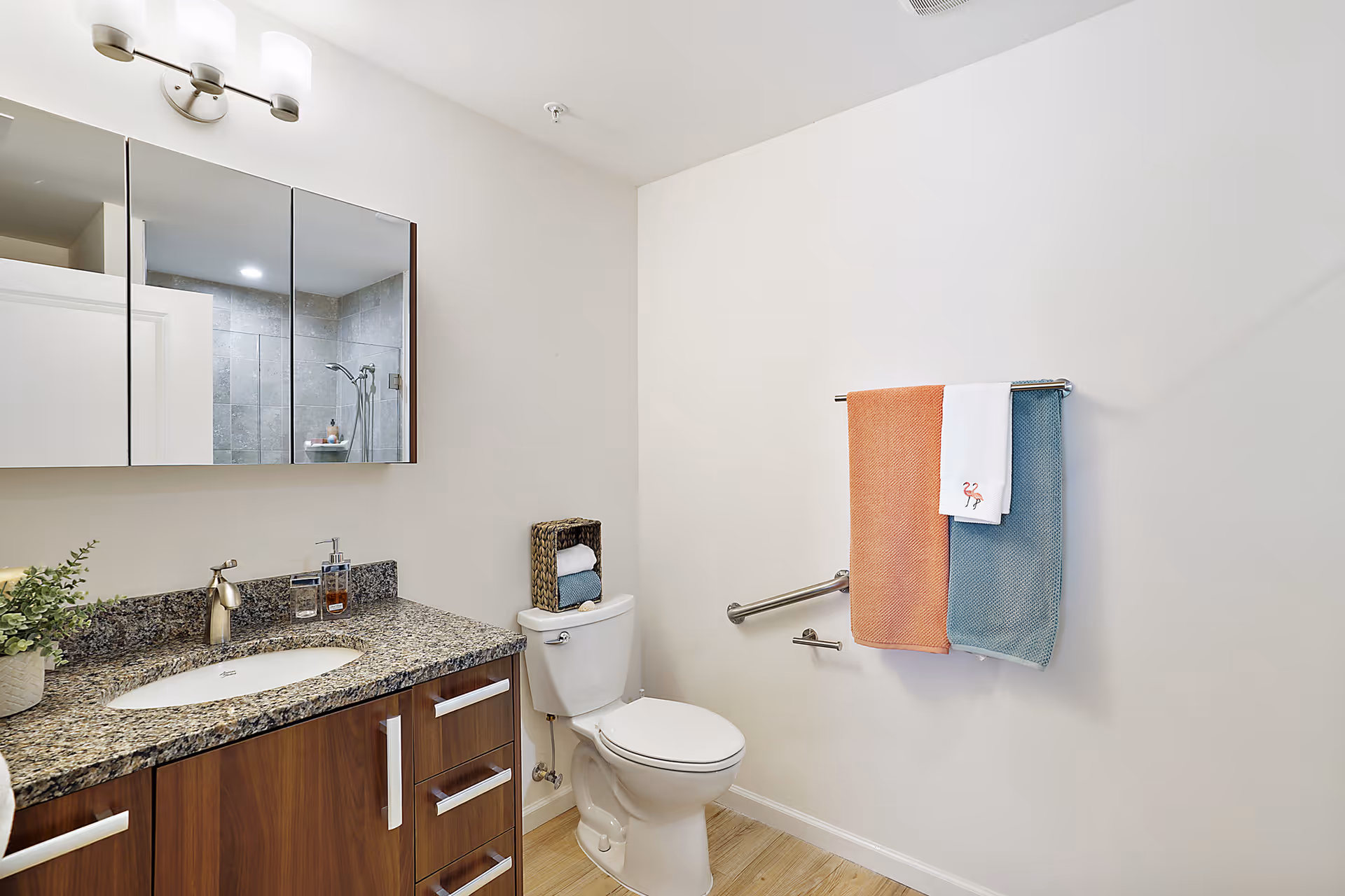 A clean and modern bathroom featuring a granite countertop with a sink, a wooden vanity with drawers, a toilet with a basket holding rolled towels on top, a towel rack with three towels in orange, white, and blue, and a glass-enclosed shower with tiled walls reflected in a mirrored medicine cabinet above the sink.