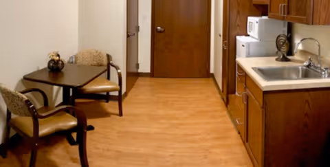 A small kitchenette area with wooden cabinets, a sink, microwave, and a small dining table with two chairs. The floor is wooden, and there is a closed wooden door at the end of the room.