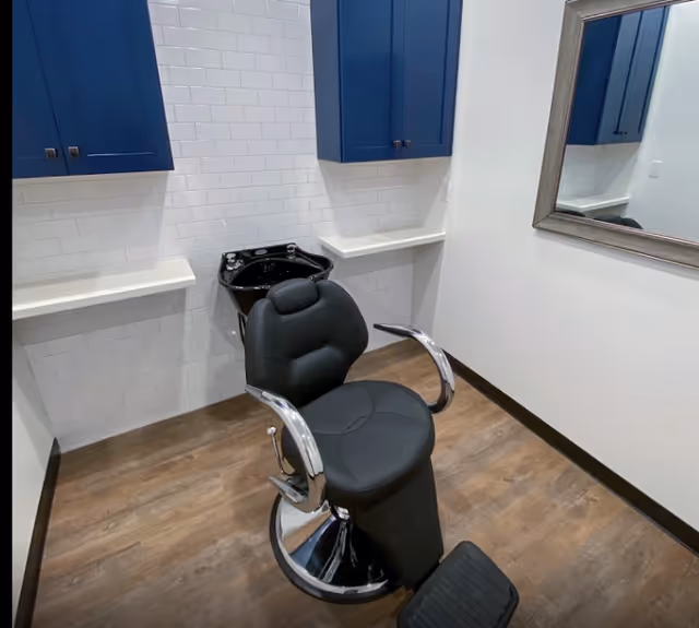A black salon chair in front of a shampoo sink in a small grooming room with blue cabinets, white tiled wall, a mirror, and wood flooring.