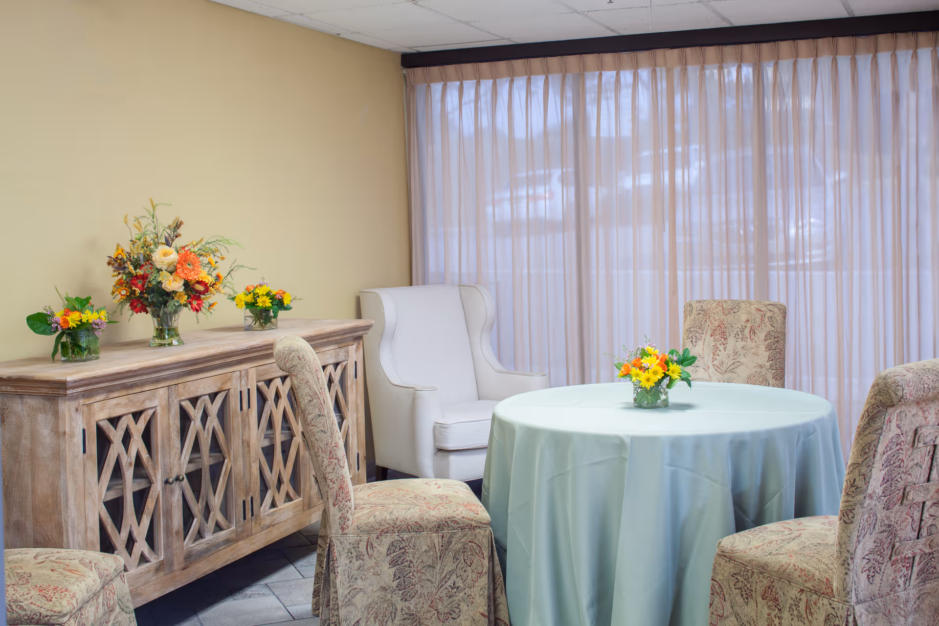 A cozy sitting area with a round table covered with a light blue tablecloth, surrounded by four upholstered chairs with a floral pattern. Behind the table is a wooden cabinet with glass-paneled doors, decorated with three vases of colorful flowers. A white wingback chair is placed near a large window with sheer curtains allowing natural light to filter in.