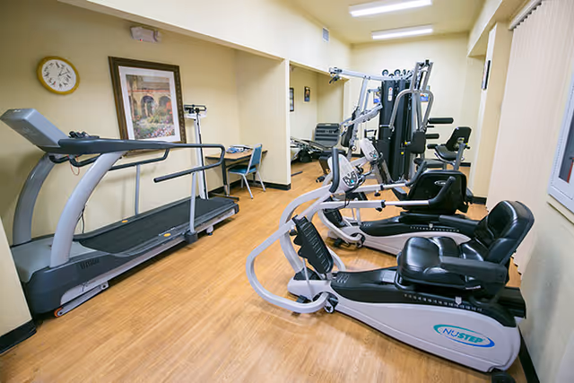A small fitness room with exercise equipment including a treadmill, a recumbent bike, and a multi-station gym machine. The room has light yellow walls, wood flooring, a clock, and framed artwork on the wall. There is also a small desk with a chair in the corner.