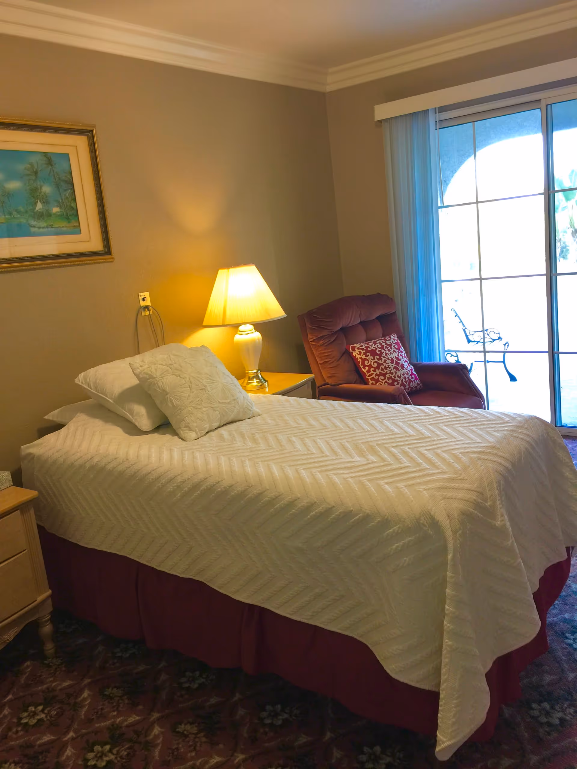A cozy bedroom with a single bed covered in a white textured bedspread and two pillows. Next to the bed is a wooden nightstand with a lit table lamp. A comfortable upholstered armchair with a decorative pillow is placed near a sliding glass door that leads to an outdoor area. A framed painting hangs on the wall above the nightstand.
