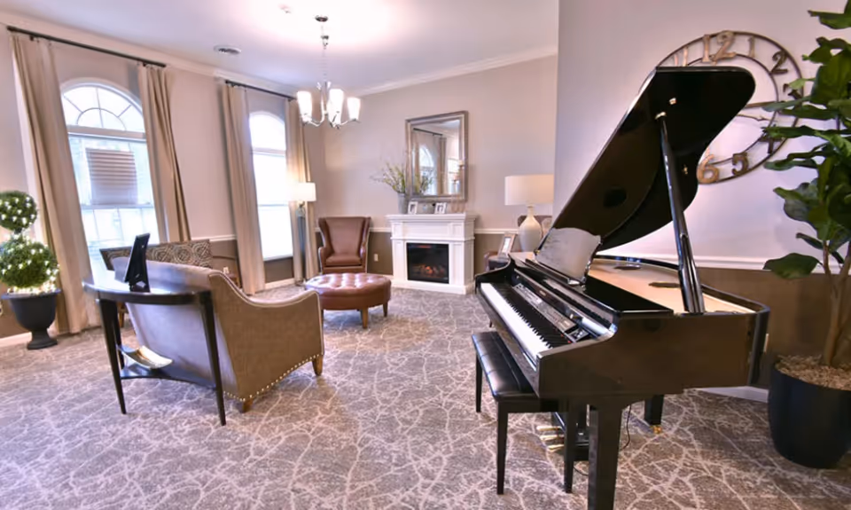 A cozy living room area with a grand piano on the right, a round ottoman, an armchair, a loveseat, and a fireplace with a mirror above it. The room has large windows with beige curtains, a chandelier, and a large wall clock. There are also decorative plants in the corners.