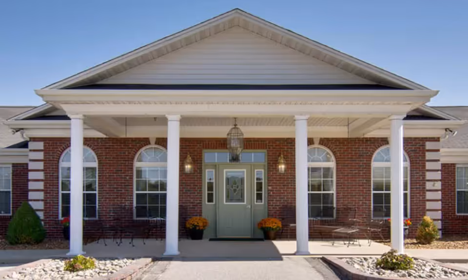 Front exterior view of a single-story brick building with a covered entrance supported by white columns. The entrance door is green with decorative glass panels, flanked by two wall-mounted lantern lights and potted plants with orange flowers. The building has arched windows with white trim and a clear blue sky above.