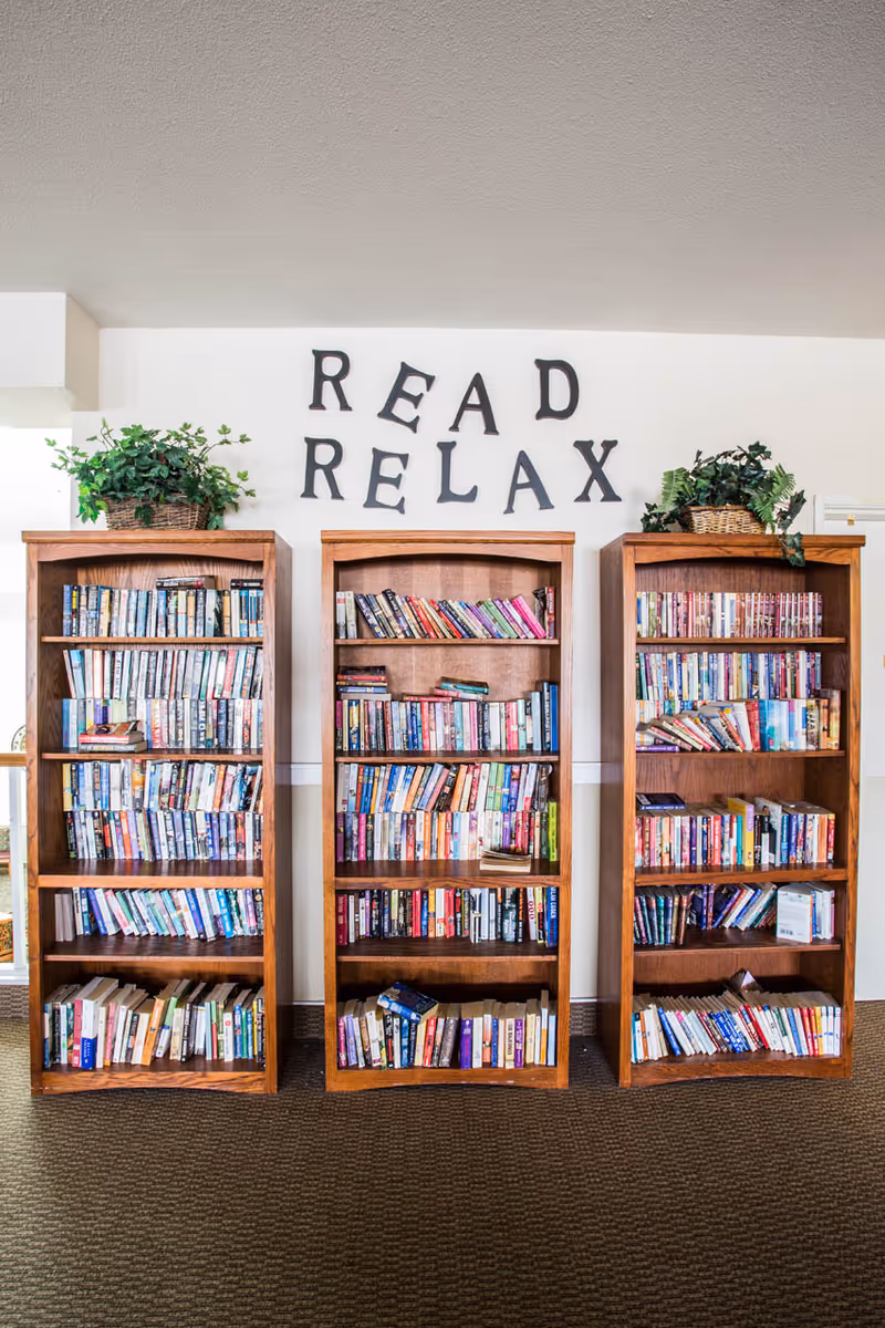 Three wooden bookshelves filled with various books arranged in a room. Above the bookshelves, black letters on the wall spell out 'READ RELAX'. Each bookshelf has a green potted plant on top. The floor is carpeted and the walls are white.