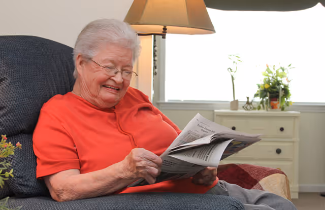 An elderly woman with white hair and glasses sitting comfortably in a cushioned chair, smiling while reading a newspaper. Behind her is a window with light coming through, a lamp, and a small dresser with plants on top.