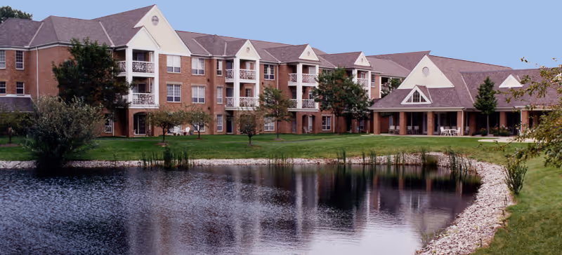 A large, multi-story brick building with balconies and a peaked roof, situated beside a pond with a rocky shoreline and surrounded by green grass and trees under a clear sky.