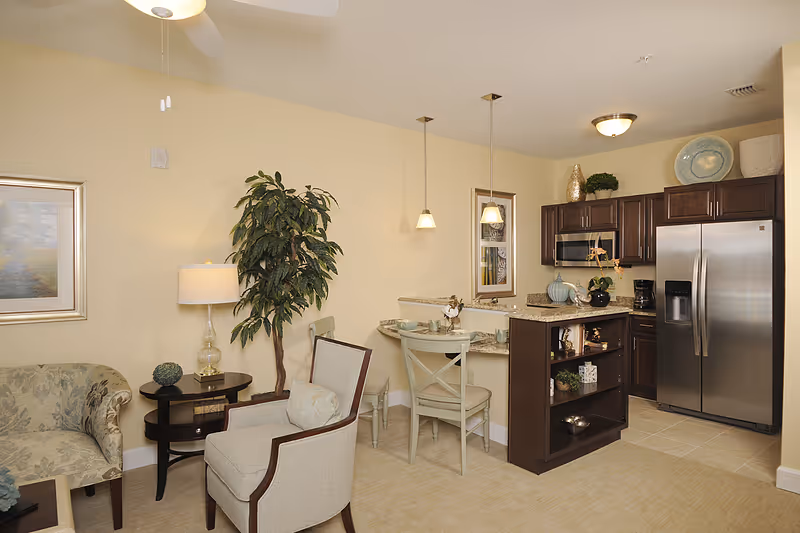 A cozy living area connected to a kitchen with dark wood cabinets and stainless steel appliances. The living area features a beige armchair, a small round side table with a lamp, a potted plant, and a patterned sofa. The kitchen includes a refrigerator, microwave, coffee maker, and a small island with shelves. There is a small dining area with two chairs and a counter with pendant lights hanging above.