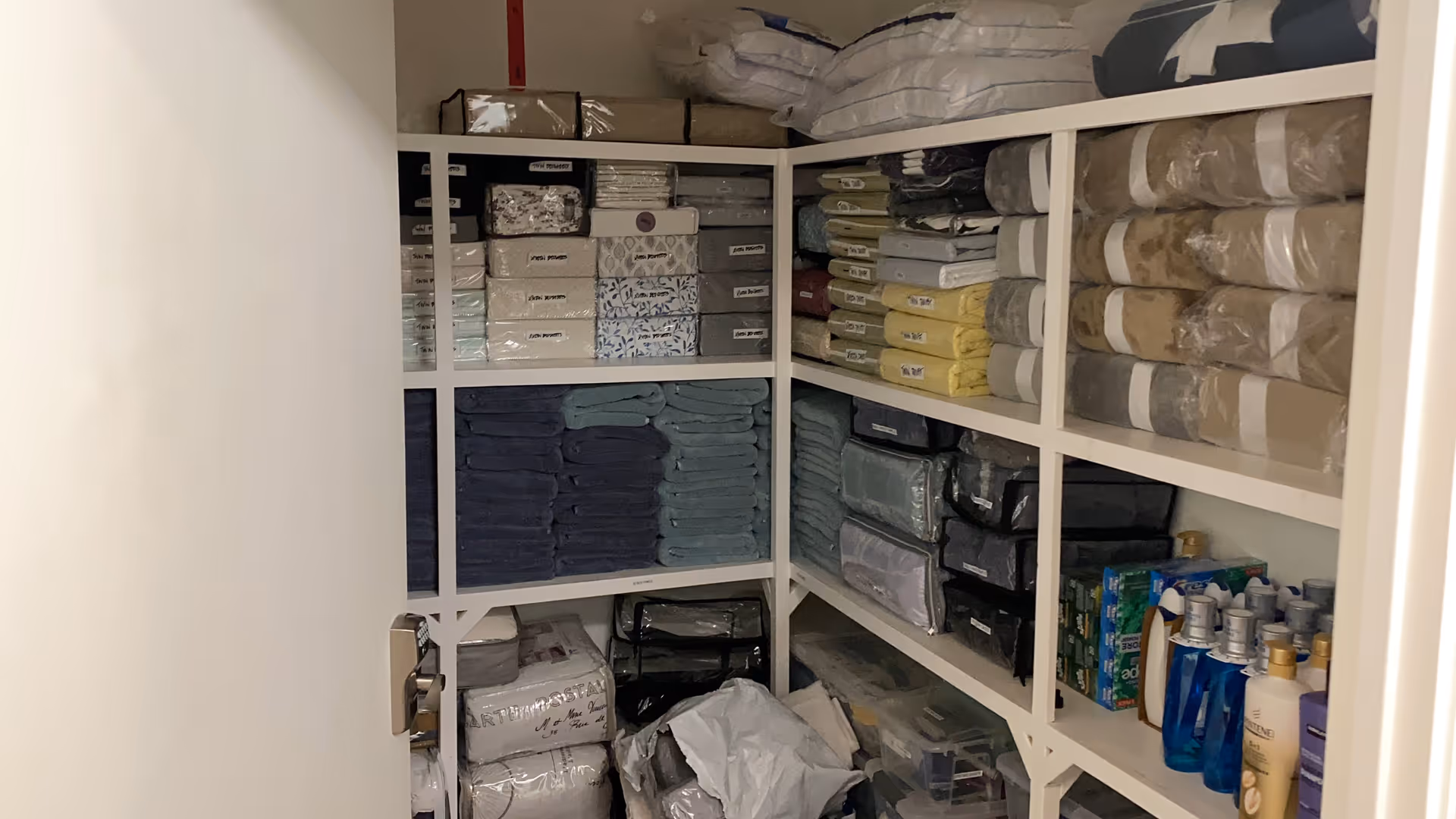 A storage closet with white shelves filled with neatly stacked towels, packaged linens, and various bottles of toiletries and cleaning supplies.