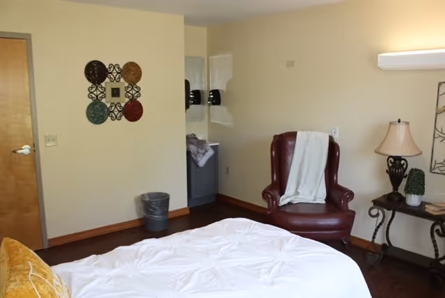 A cozy bedroom in Oakdale Care Center featuring a bed with white bedding and a yellow pillow, a burgundy leather armchair with a white blanket draped over it, a small table with a lamp and decorative items, a wall decoration with colorful circular plates, a trash can, and a small sink area in the corner.
