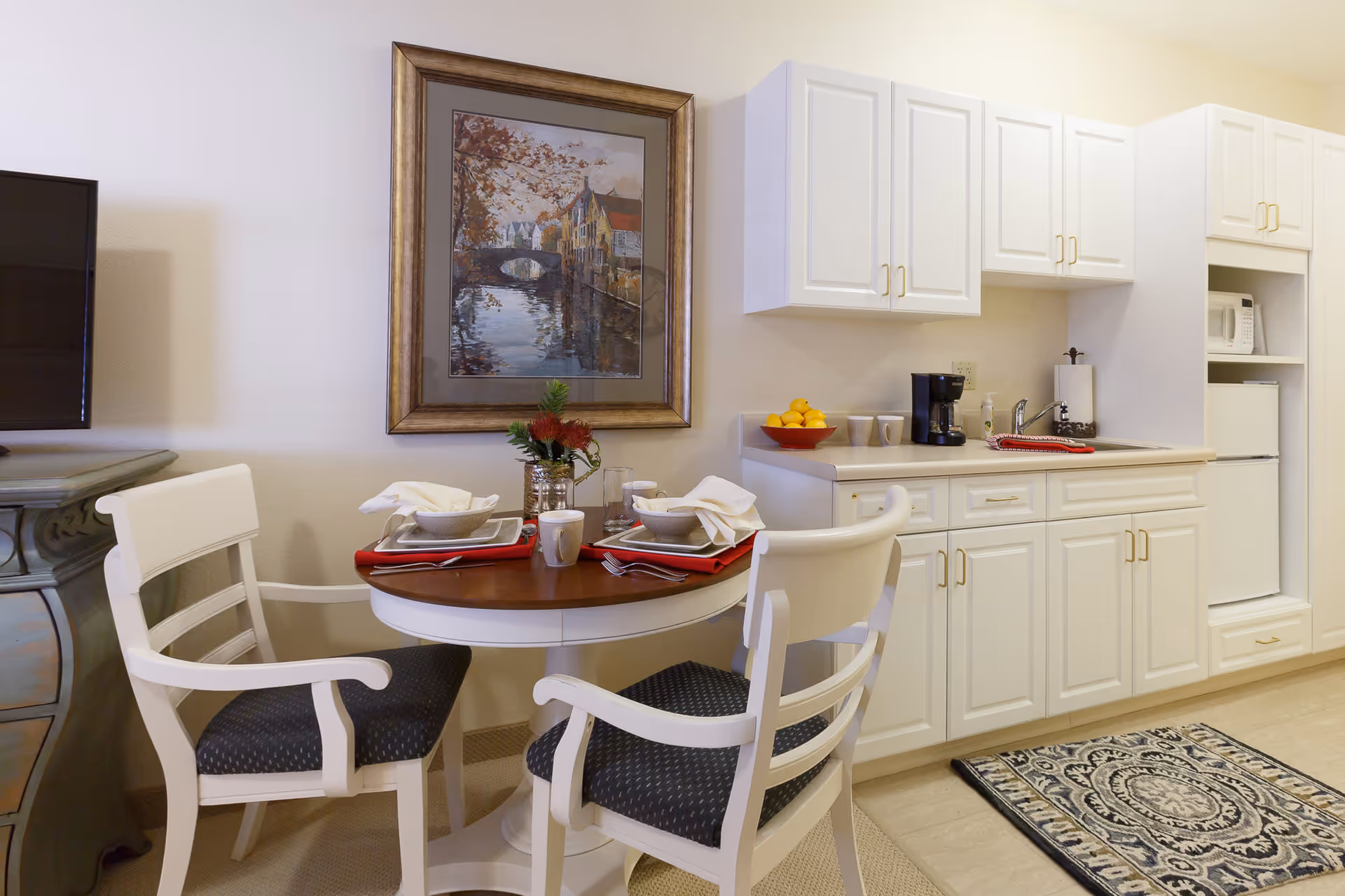 A small dining area with a round wooden table set for two with white chairs that have dark cushioned seats. The table is set with bowls, plates, mugs, and napkins. Behind the table is a kitchen area with white cabinets, a coffee maker, a bowl of lemons, a microwave, and a small refrigerator. A framed painting of a canal scene hangs on the wall above the table. A patterned rug is on the floor in front of the kitchen cabinets.