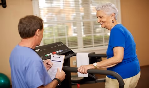 An elderly woman in a blue shirt is walking on a treadmill while a man in a blue medical scrub is holding a clipboard and observing her in a well-lit room with large windows.