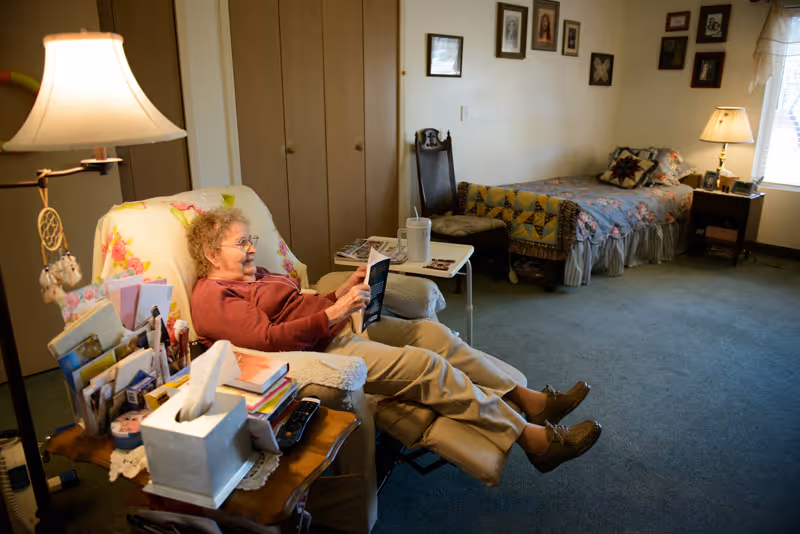 An elderly woman reclining in a cushioned armchair reading a book in a cozy room. The room has a single bed with a quilt, a wooden chair, a side table with a lamp, and framed pictures on the wall. There is a window letting in natural light and a carpeted floor.