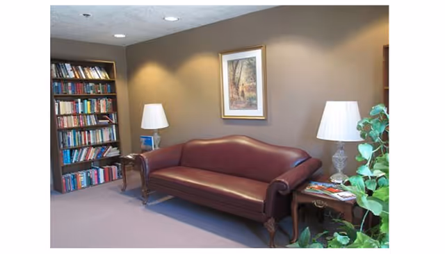 A small residential common room with a burgundy leather sofa flanked by side tables and lamps, a bookshelf, framed artwork, and a potted plant.