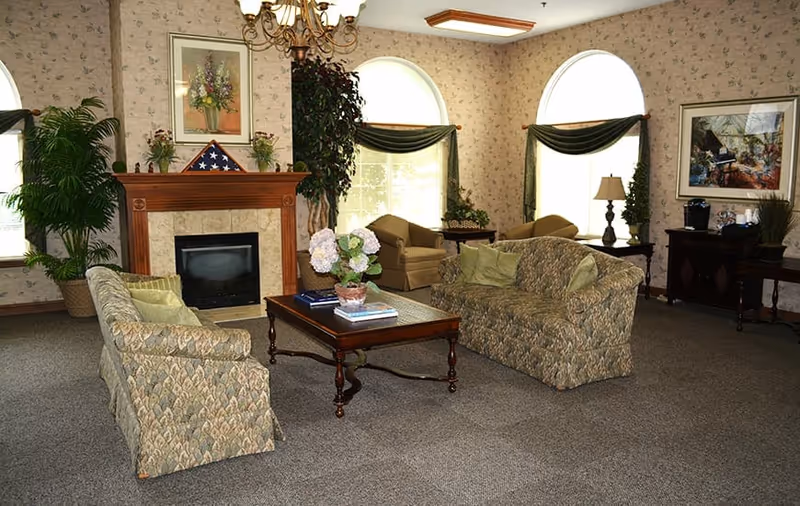 A cozy living room with two patterned sofas facing each other around a wooden coffee table with a flower pot and books. There is a fireplace with a folded American flag on the mantel, two large arched windows with green curtains, a chandelier, and framed artwork on the walls. Additional seating and a side table with a lamp are visible near the windows.