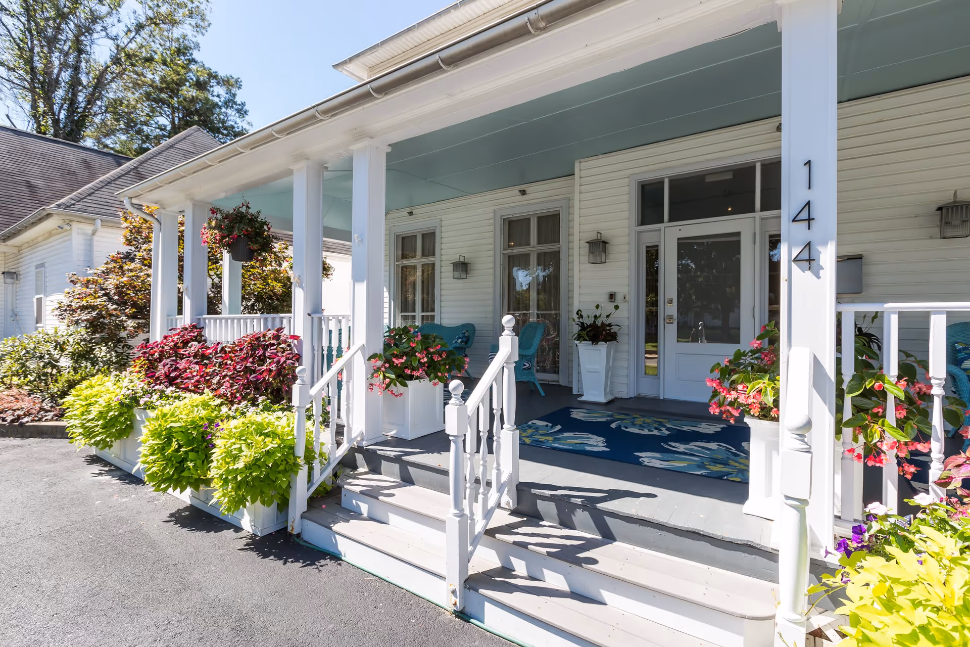 Front porch of a white senior living facility with steps leading up to a covered entrance. The porch is decorated with potted plants and flowers, blue chairs, and a blue floral rug. The building number 144 is visible on a white column.