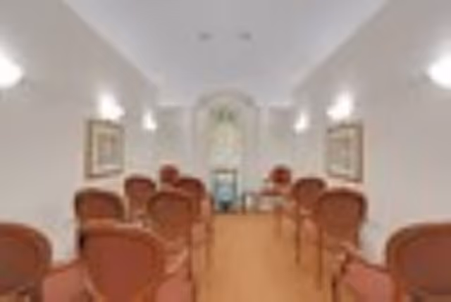 A narrow interior meeting or chapel-style room with rows of red upholstered chairs facing an arched decorative window at the far end.