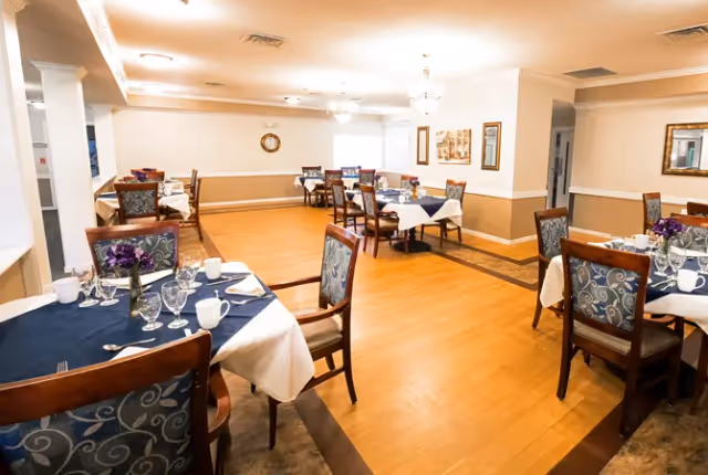 A bright dining room with several tables covered in white and navy blue tablecloths, set with glasses, cups, and silverware. The chairs have patterned upholstery, and the room features light-colored walls with decorative trim, a clock, framed artwork, and ceiling lights.