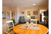 Bright assisted-living common area with a round wooden dining table set with newspapers and chairs, and upholstered armchairs, lamp, and TV in the background.
