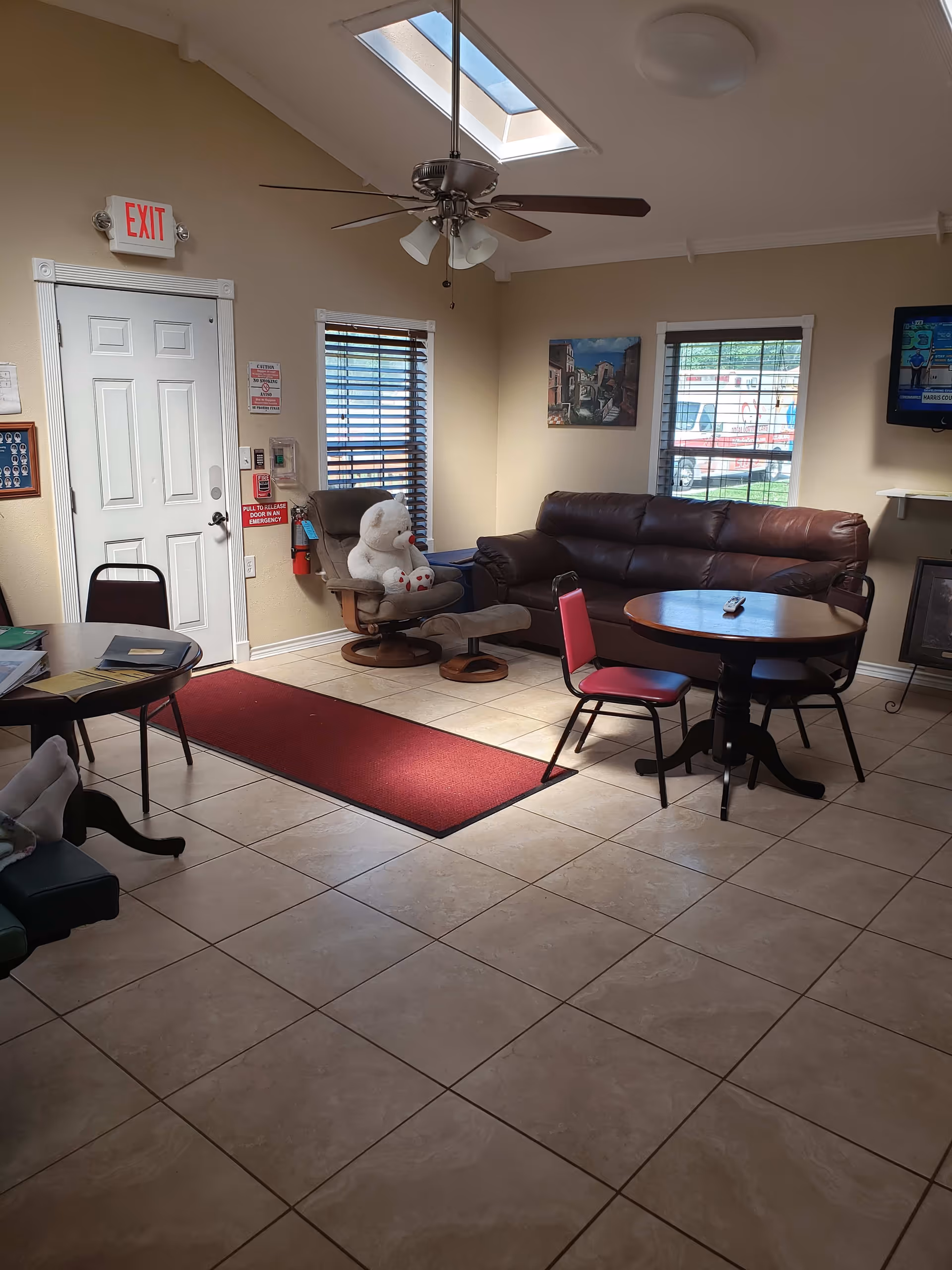 A cozy living room area with beige tiled floor, a brown leather couch, a round wooden table with three chairs, a reclining chair with a large white teddy bear on it, two windows with blinds, a ceiling fan with lights, a skylight, a wall-mounted TV, and an exit door with an illuminated exit sign above it.