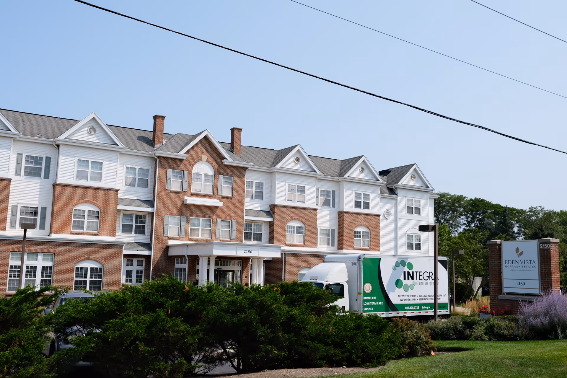 Front exterior of the three-story brick-and-white Eden Vista Hoffman Estates senior living building with landscaping and a parked service truck.