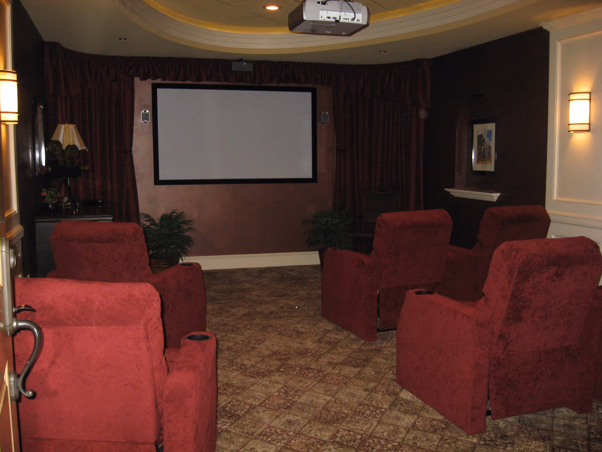 A small home theater room with six red upholstered recliner chairs arranged in two rows facing a large blank projection screen. The room has patterned carpet flooring, dark red curtains on the walls, two potted plants near the screen, wall sconces providing soft lighting, and a ceiling-mounted projector.