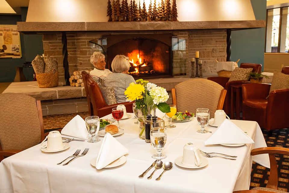 A table set for four with white linens, glasses, and a floral centerpiece in a cozy dining room with a lit fireplace and two seated seniors in the background.