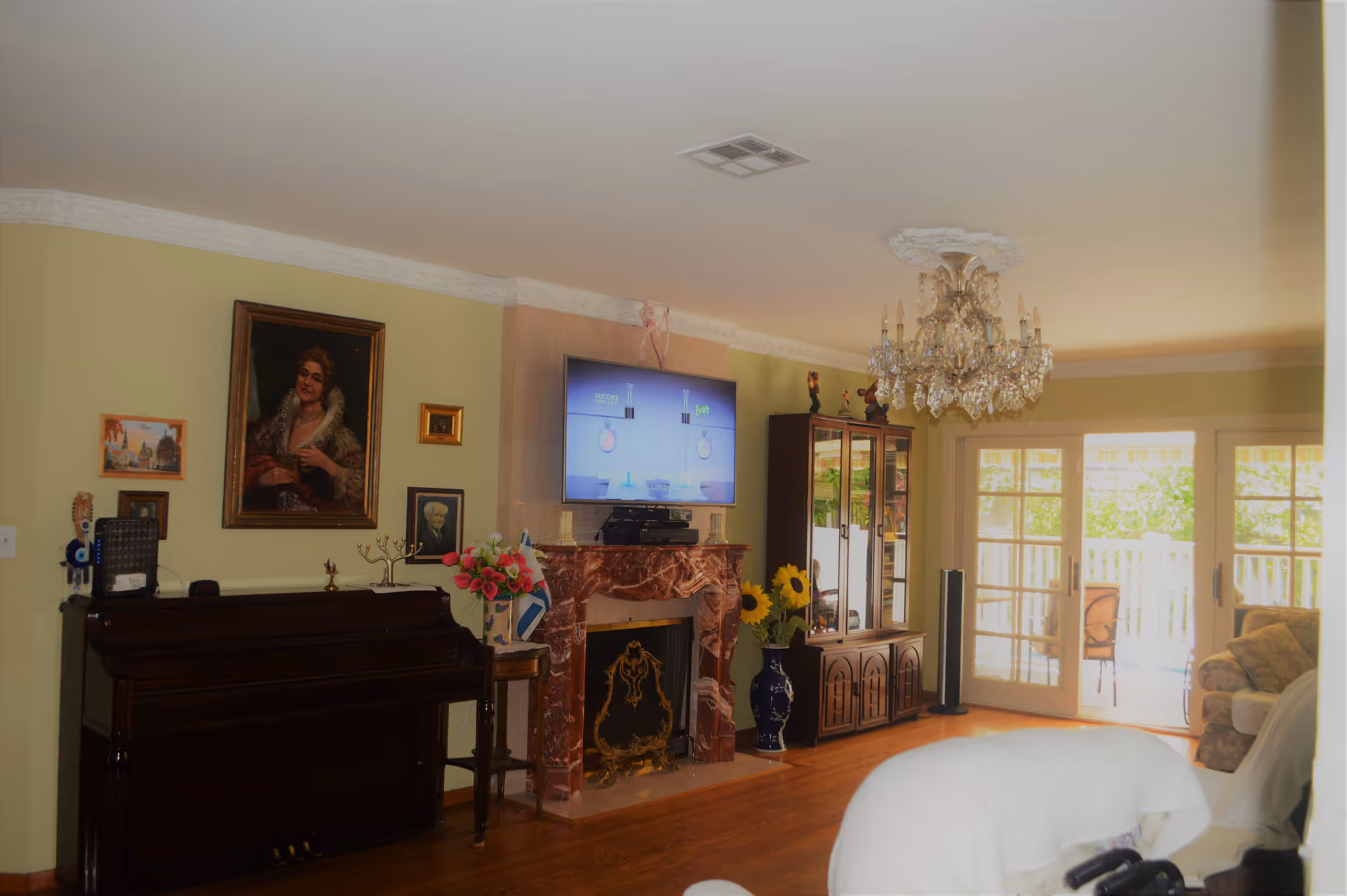 A cozy living room with a chandelier hanging from the ceiling, a flat-screen TV mounted above a decorative red marble fireplace, a dark wooden piano against the wall with framed artwork above it, a glass-front cabinet with figurines, a vase with sunflowers, and sliding glass doors leading to an outdoor patio with chairs.