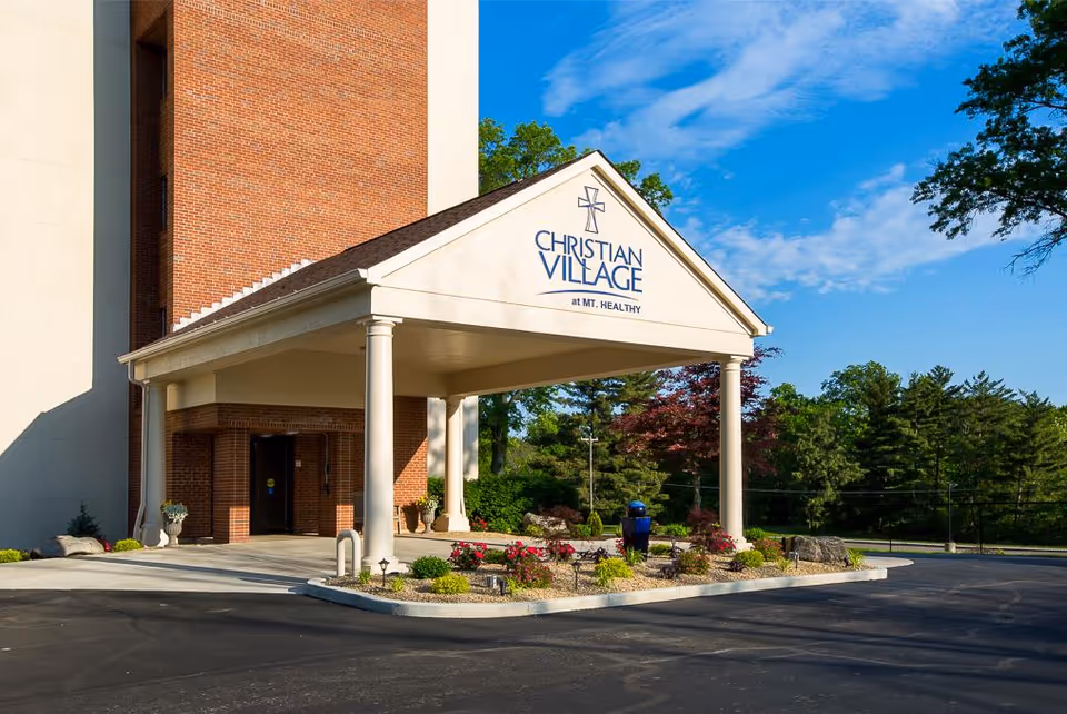 Entrance of a brick building with a covered driveway supported by four white columns. The sign on the front of the covered entrance reads 'Christian Village at Mt. Healthy'. There are landscaped flower beds with shrubs and flowers in front of the entrance. Trees and a blue sky with some clouds are visible in the background.