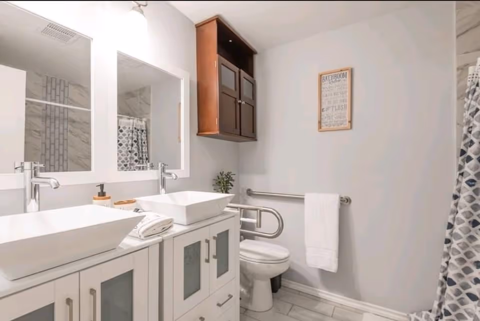 Well-lit bathroom with double vessel sinks and mirrors, a toilet with a safety grab bar, and a patterned shower curtain.