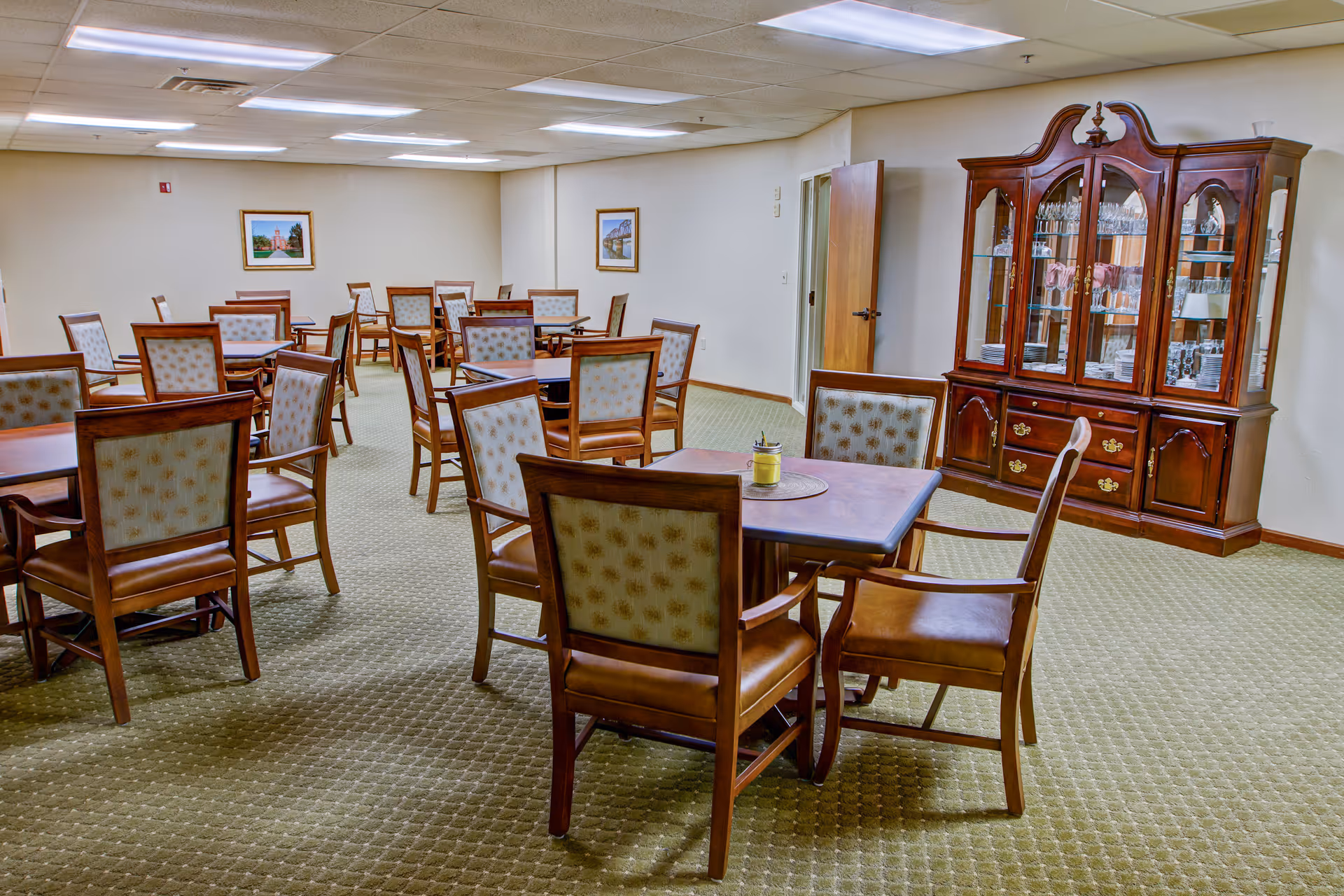 Empty communal dining room with multiple wooden tables and upholstered chairs and a glass-front china cabinet.