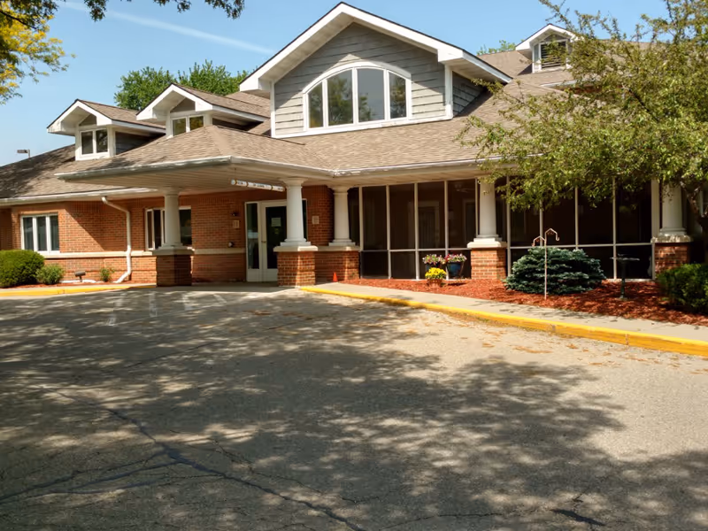 Front entrance of a brick-and-siding retirement center with a covered porte-cochere, screened porch, and landscaped beds.