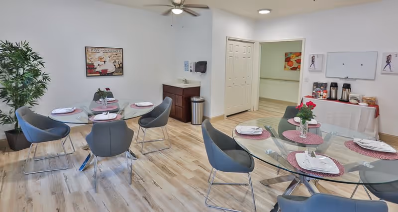 Bright communal dining area with round glass tables set for meals, gray chairs, a beverage station, and light wood flooring.