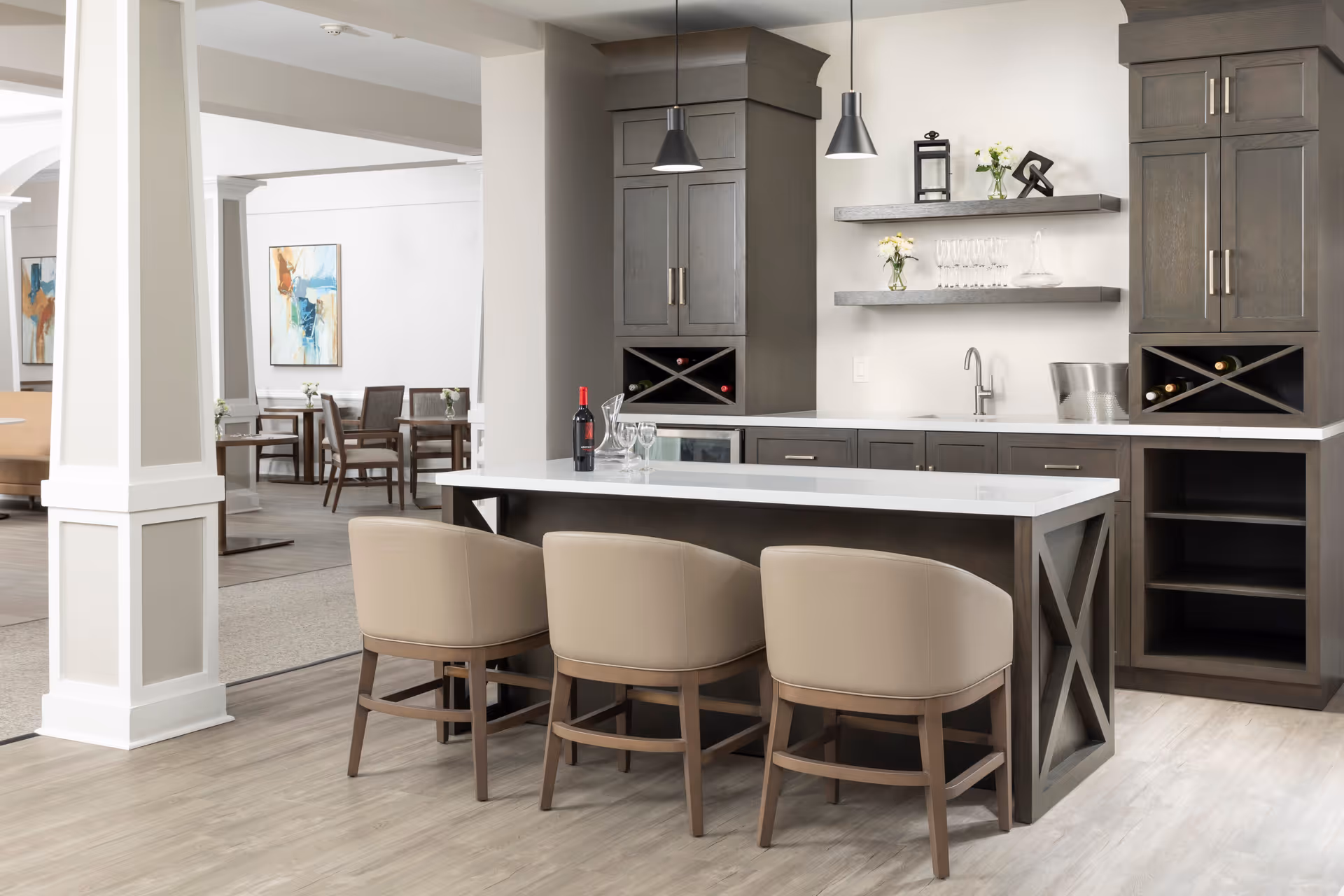 Modern kitchen area in an assisted living facility with a white countertop island, three beige cushioned chairs, dark wood cabinets, wine racks, a small sink, and shelves with glassware and decorative items. In the background, there is a dining area with tables, chairs, and abstract wall art.