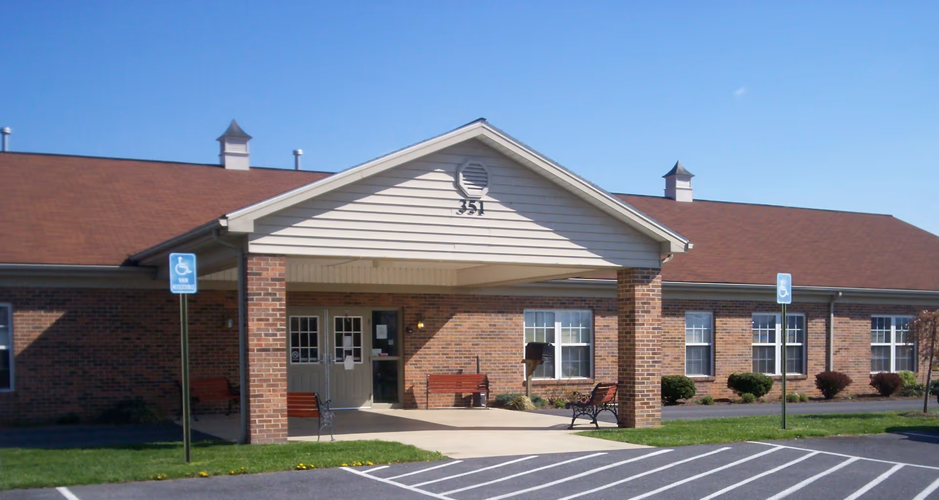 Front entrance of a single-story brick senior living building with a covered portico, benches, and marked handicap parking spaces.