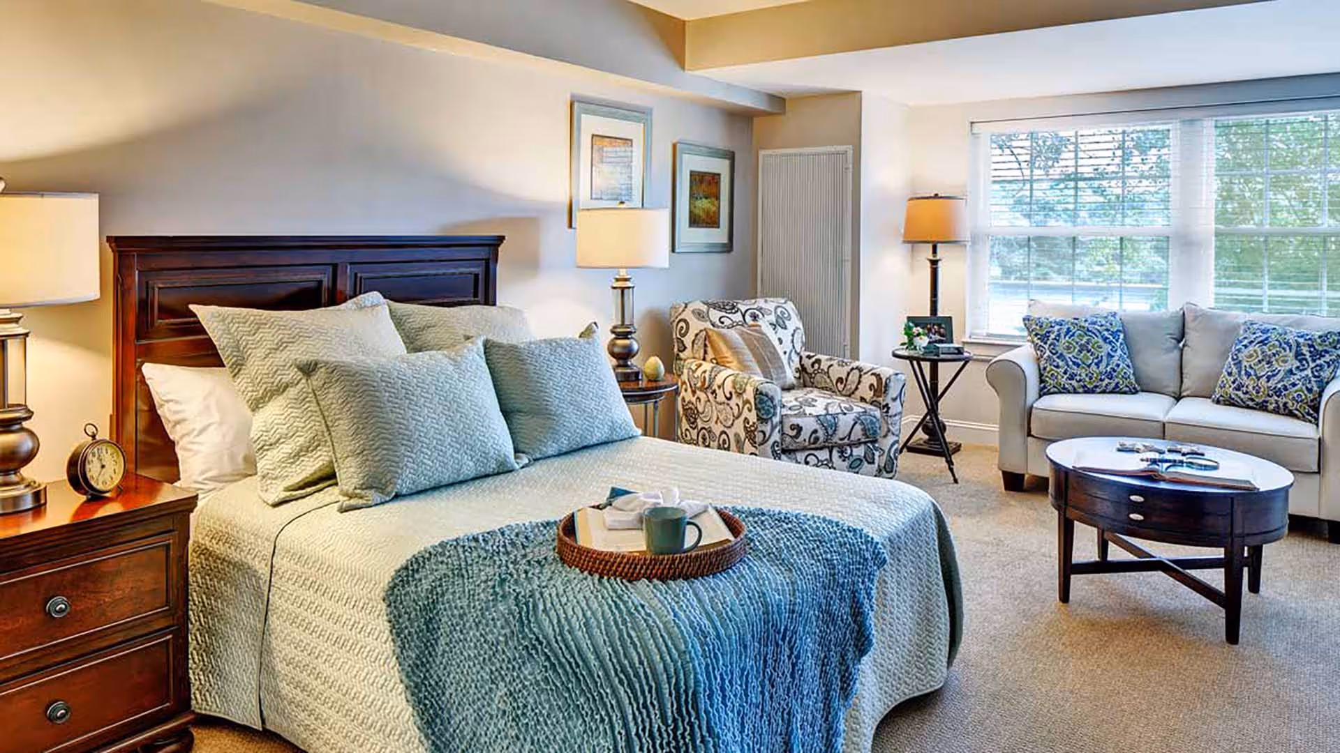 A cozy bedroom with a neatly made bed featuring multiple pillows and a blue throw blanket. There is a wooden nightstand with a lamp and an alarm clock on the left side of the bed. In the background, there is a patterned armchair, a small round table with a lamp, and a sofa with decorative pillows near a large window letting in natural light.
