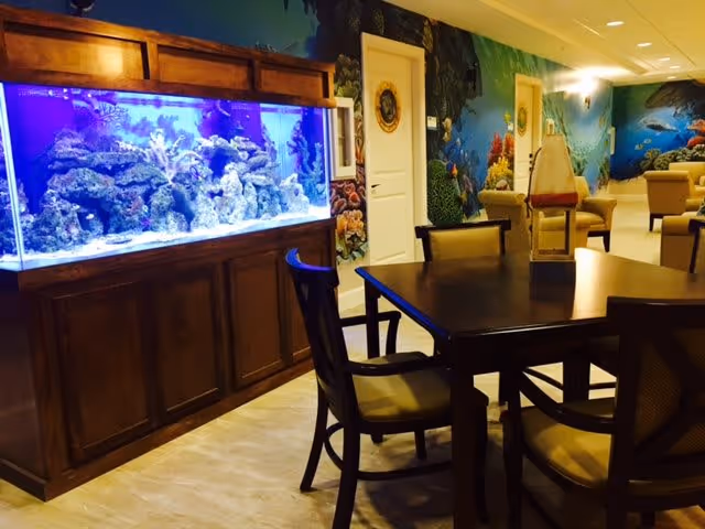 Interior view of a common area featuring a large built-in aquarium filled with coral and fish on the left, a dark wooden table with cushioned chairs in the foreground, and walls decorated with colorful underwater-themed murals.